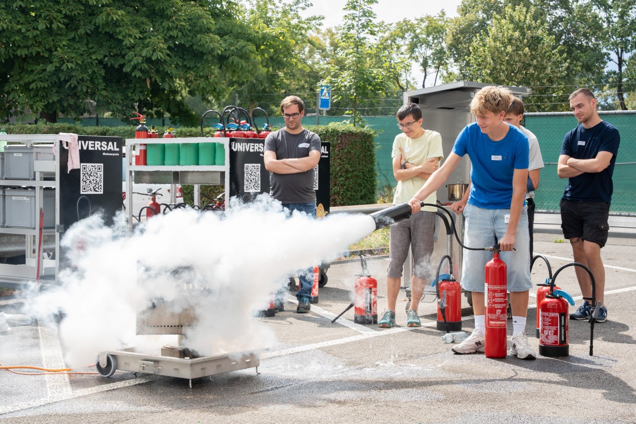 Ein Jugendlicher löscht ein Feuer mit einem Feuerlöscher und im Hintergrund sehen ihn dabei weitere Jugendliche zu.