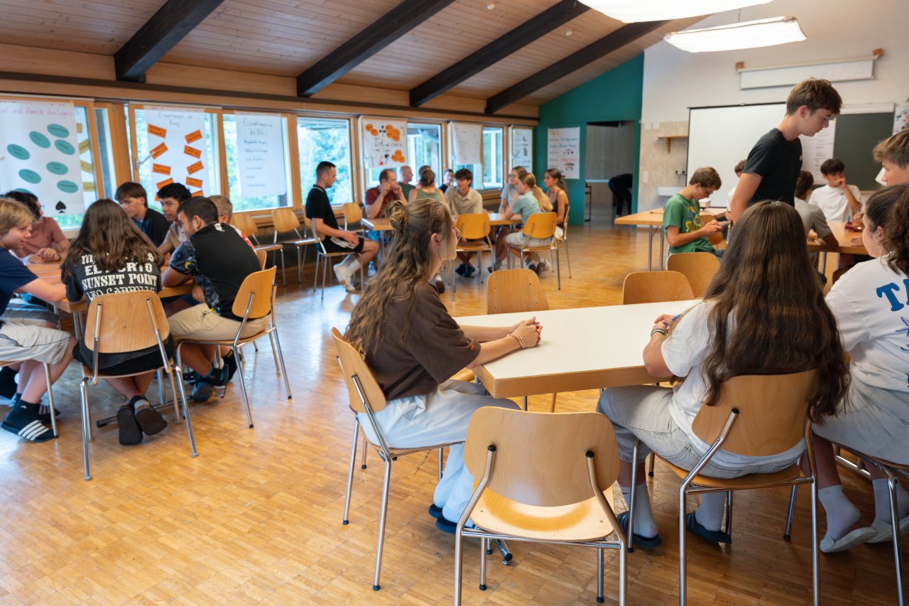 Eine Gruppe Jugendlicher sitzen in einem Schulraum in verschiedenen Gruppen zusammen und lösen Aufgaben.
