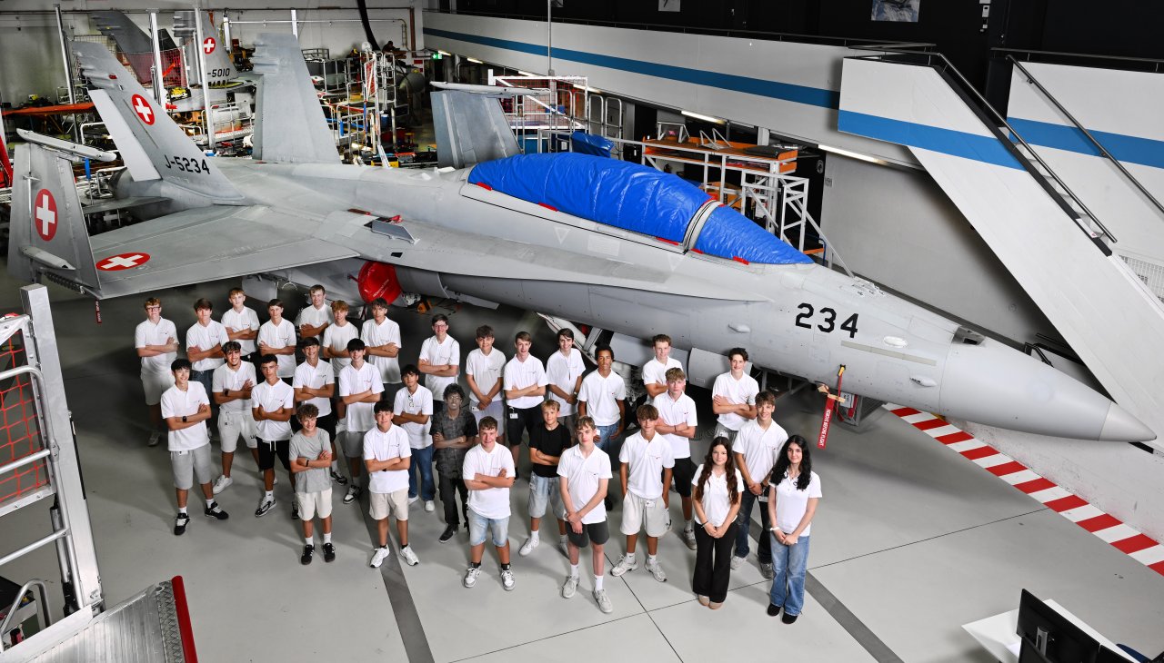 Eine Gruppe von jungen Menschen stehen in einer Halle vor einem FA-18 Kampfjet und werden von oben fotografiert.