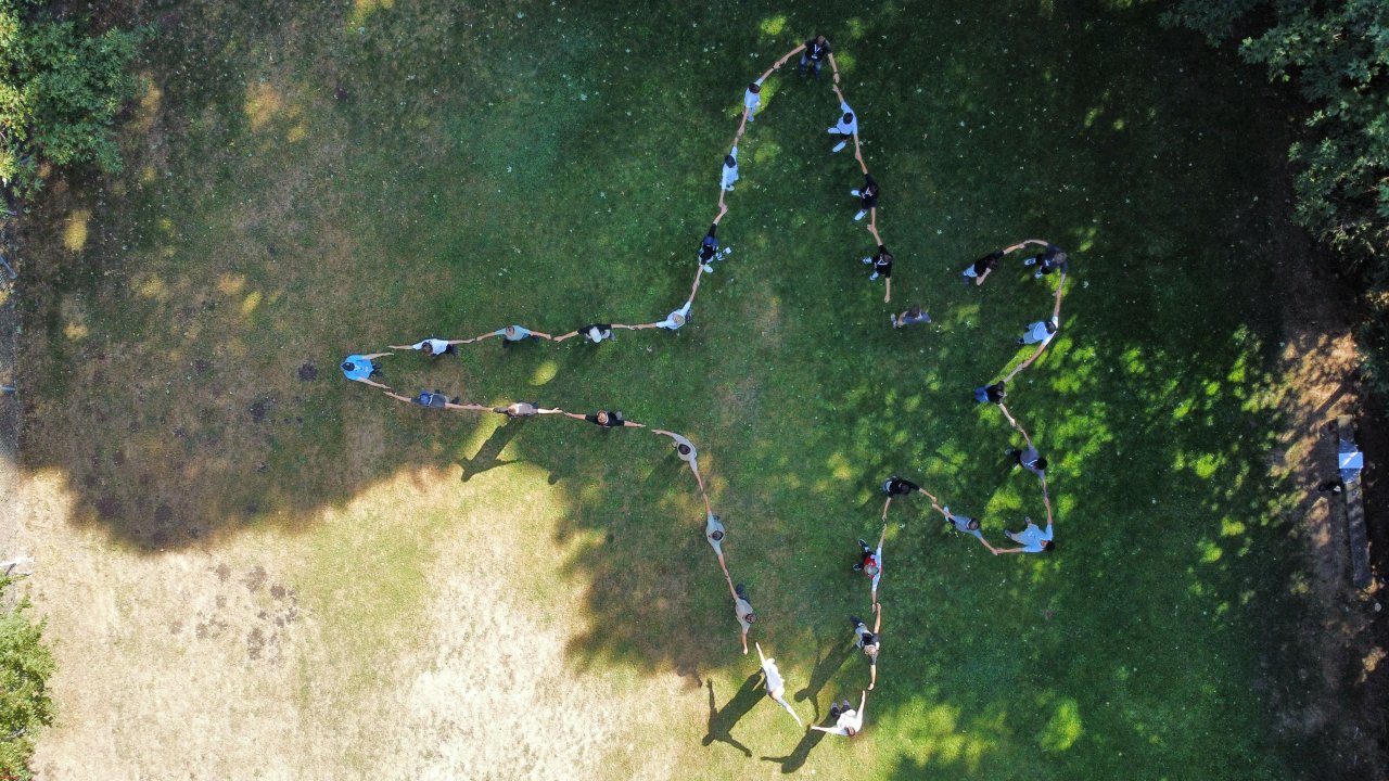 Eine Gruppe von Jugendlichen liegen auf einer grünen Wiese und formen mit ihren Körper einen Umriss von einem Kampfjet.
