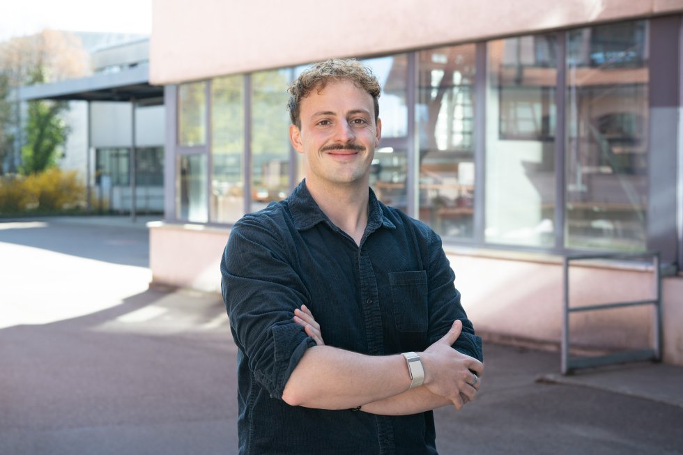 Ein junger Mann in dunkler Kleidung steht vor einem Gebäude mit verschränkten Armen.